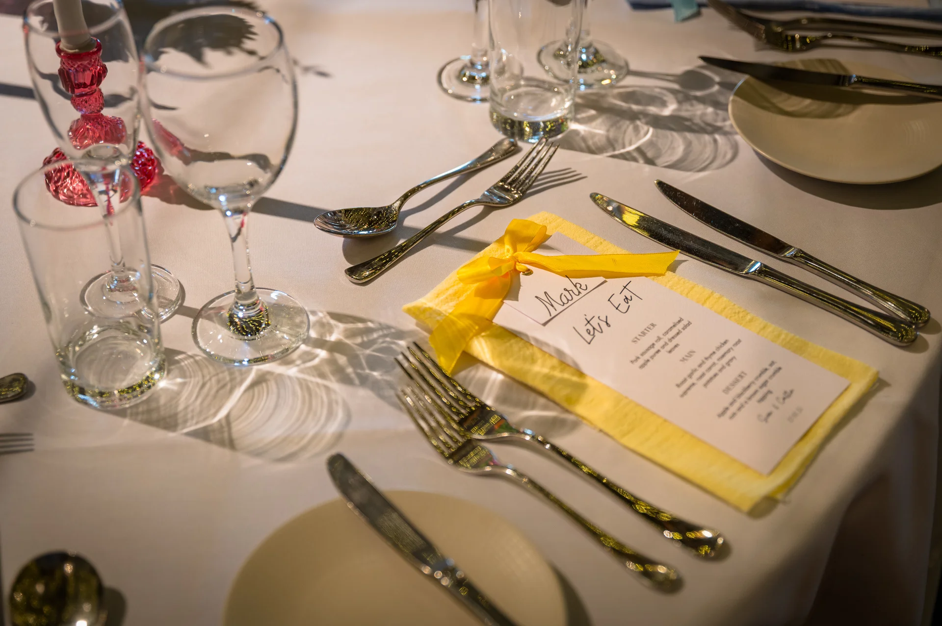A beautifully styled wedding tablescape features yellow napkins tied with bright yellow bows, custom menus, gleaming cutlery, and crystal glassware under warm lighting.