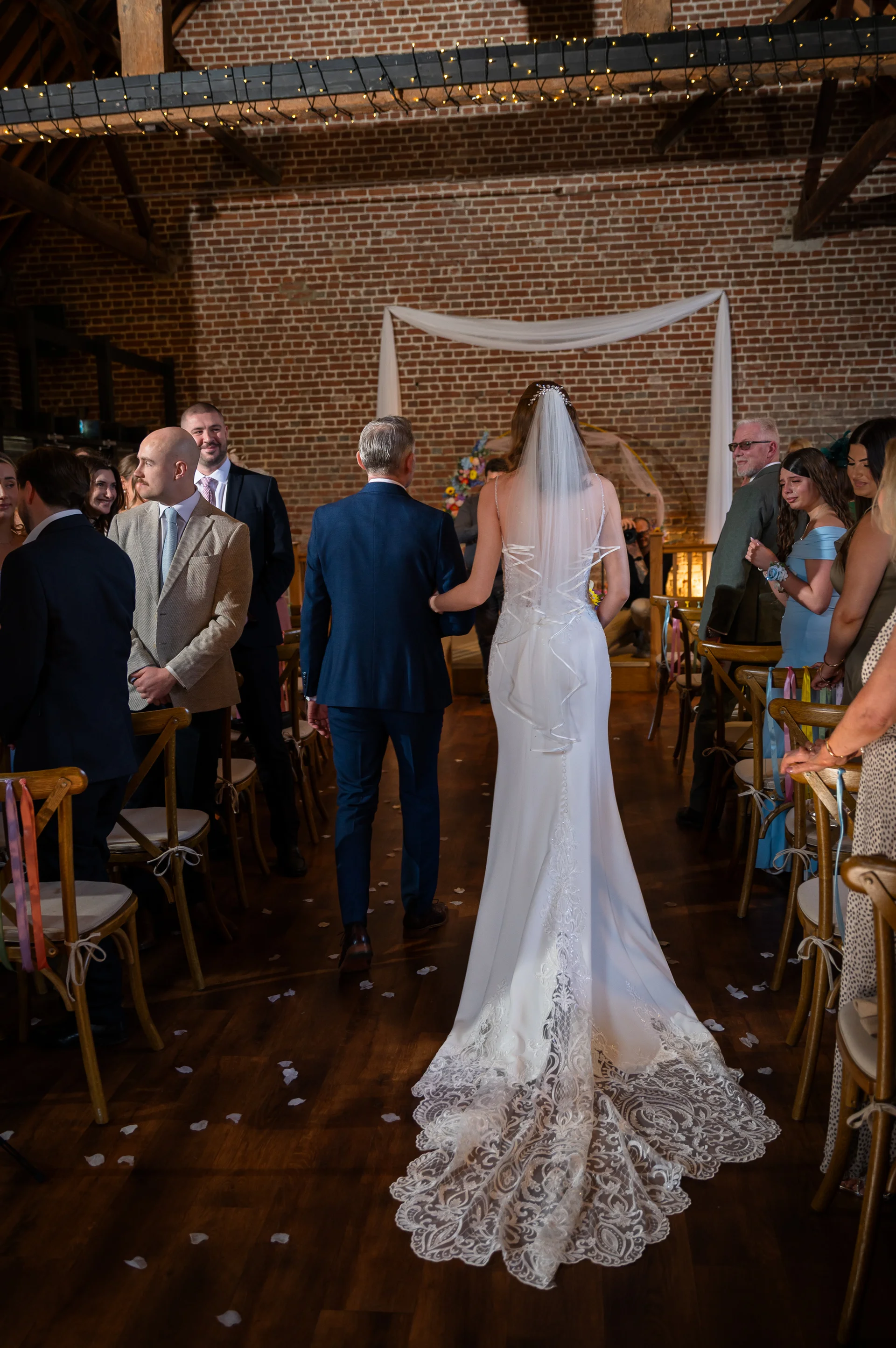 The bride and her father walk down the wooden aisle of a rustic brick barn wedding venue, showcasing her fitted white gown with a detailed lace train and veil.