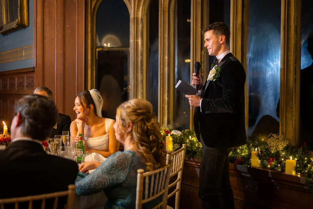 A groom in a velvet tuxedo stands with a microphone giving his wedding speech while the laughing bride looks on from the candlelit top table.