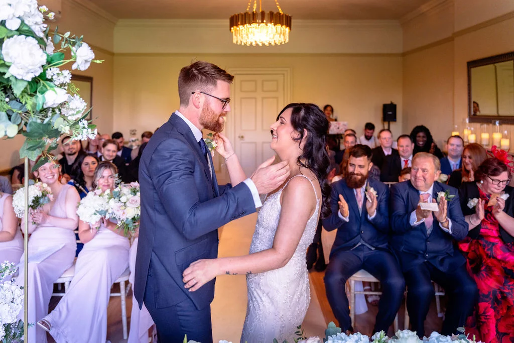 A newlywed couple smiling lovingly at each other during their wedding ceremony as seated guests applaud enthusiastically in a bright room.
