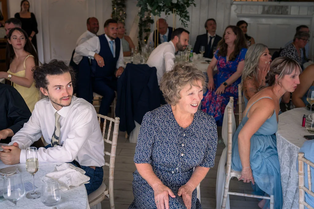 Wedding guests gather around white-clothed tables, smiling and reacting to a speech during the reception celebration.