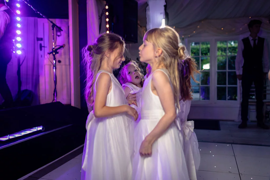 Young flower girls in white dresses dance together on an illuminated dance floor under vibrant purple reception lighting.