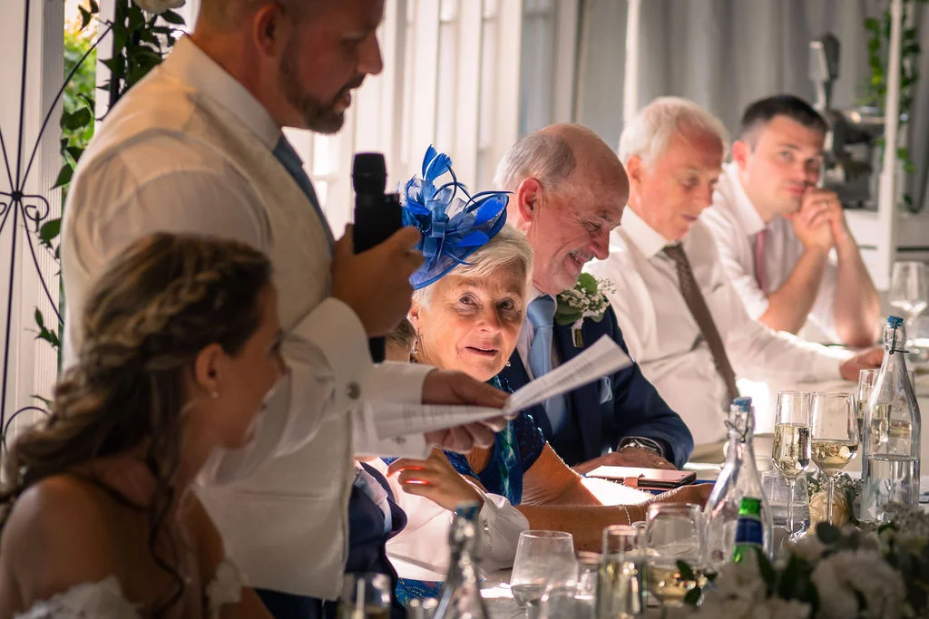 A guest delivers a heartfelt wedding speech at the top table while family members listen and smile during the reception.