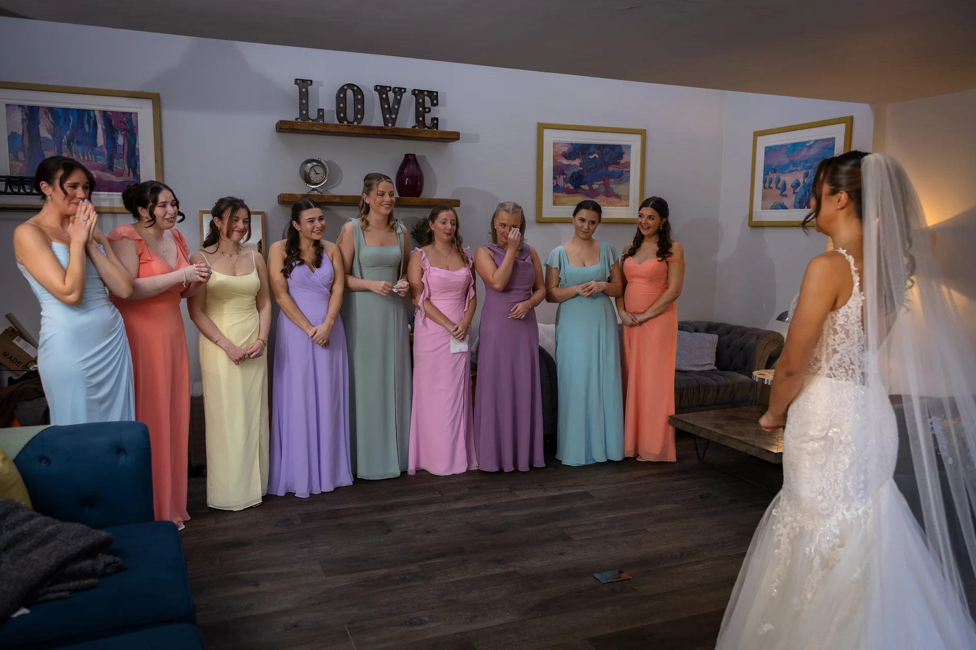 A beautiful bride in a lace wedding dress and veil reveals her look to her bridesmaids, who stand in a row wearing a colourful array of pastel dresses.