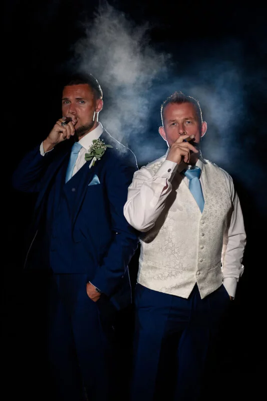 Two well-dressed groomsmen pose dramatically against a dark background while smoking cigars amidst thick plumes of smoke.