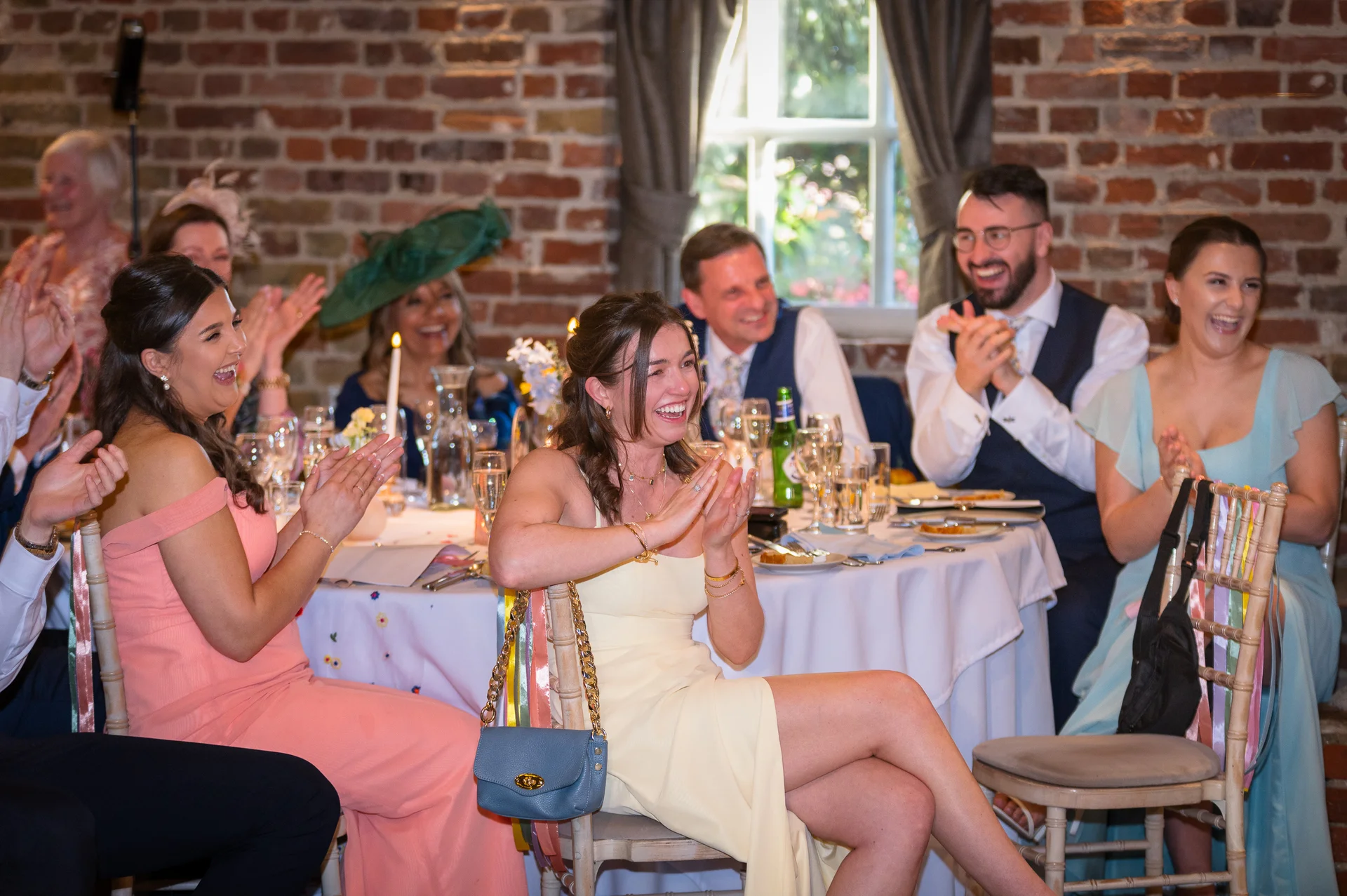 Wedding guests sitting around a round banquet table in a rustic brick venue, clapping and laughing joyfully during wedding speeches.