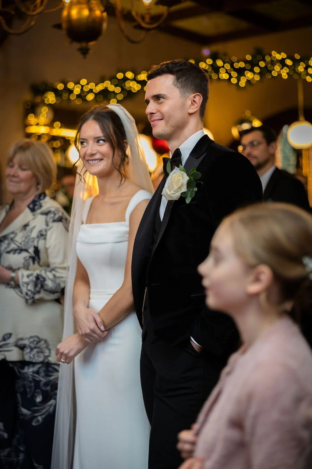 A smiling bride and groom stand together during their wedding ceremony, surrounded by guests and softly glowing fairy lights in the background.