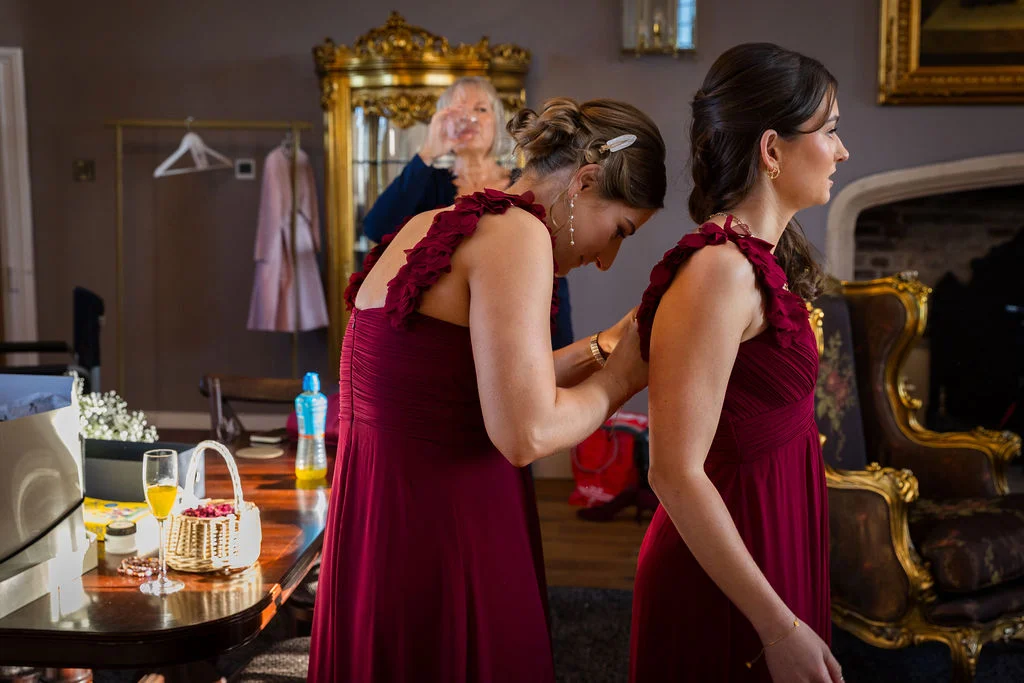 A bridesmaid in a burgundy dress helps another woman fasten her gown in a warmly lit, elegant room during morning bridal preparations.