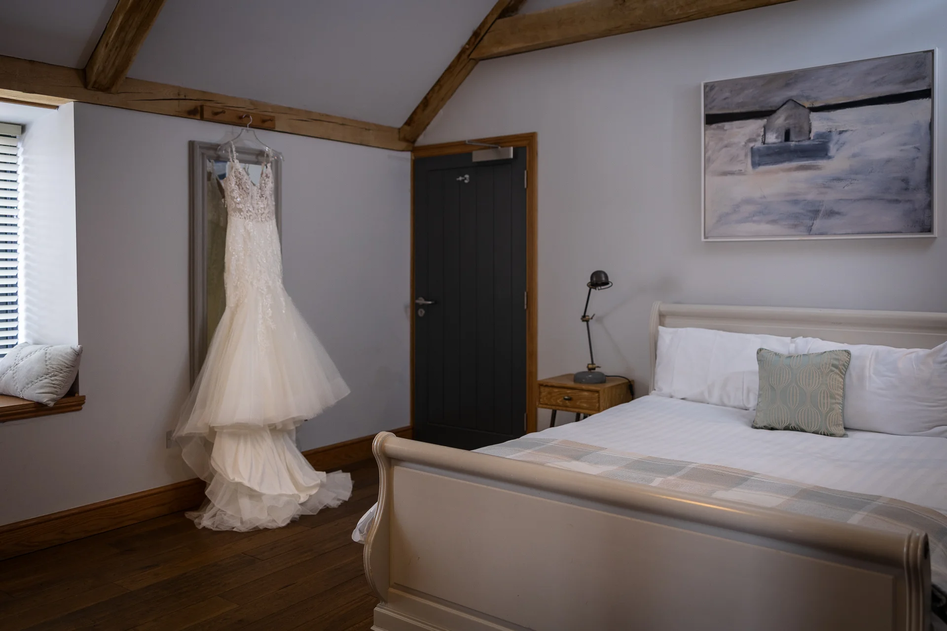 An elegant lace wedding dress hangs against a mirror in a light-filled hotel bedroom with exposed wooden beams and a made bed.