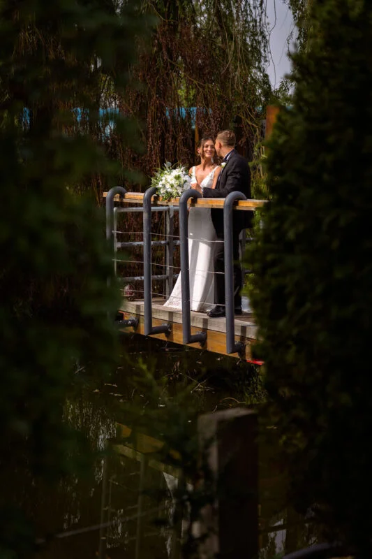 A bride and groom standing on a bridge over a pond.
