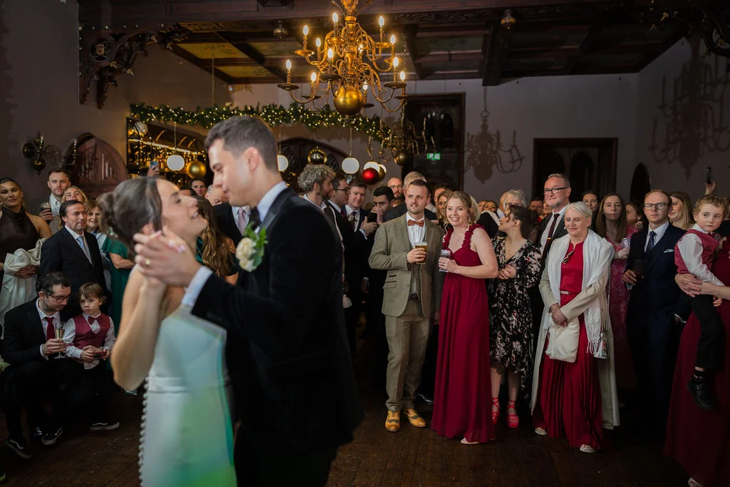 The newlywed couple share their romantic first dance in a grand, chandelier-lit hall while wedding guests look on and smile.