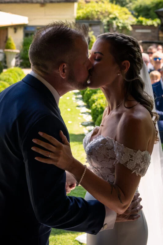 The bride and groom share their first kiss outdoors in a garden setting, illuminated by warm natural sunlight.