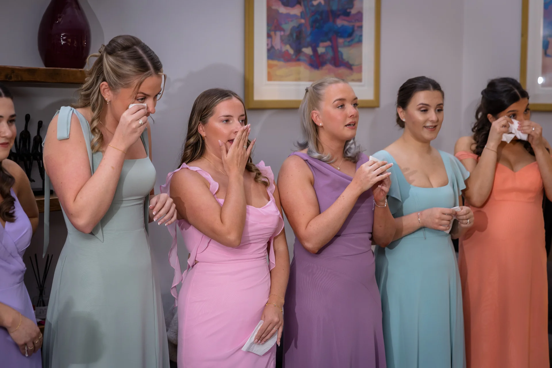 A line of emotional bridesmaids in colourful pastel dresses wipe away tears and react with moving expressions during the wedding ceremony.