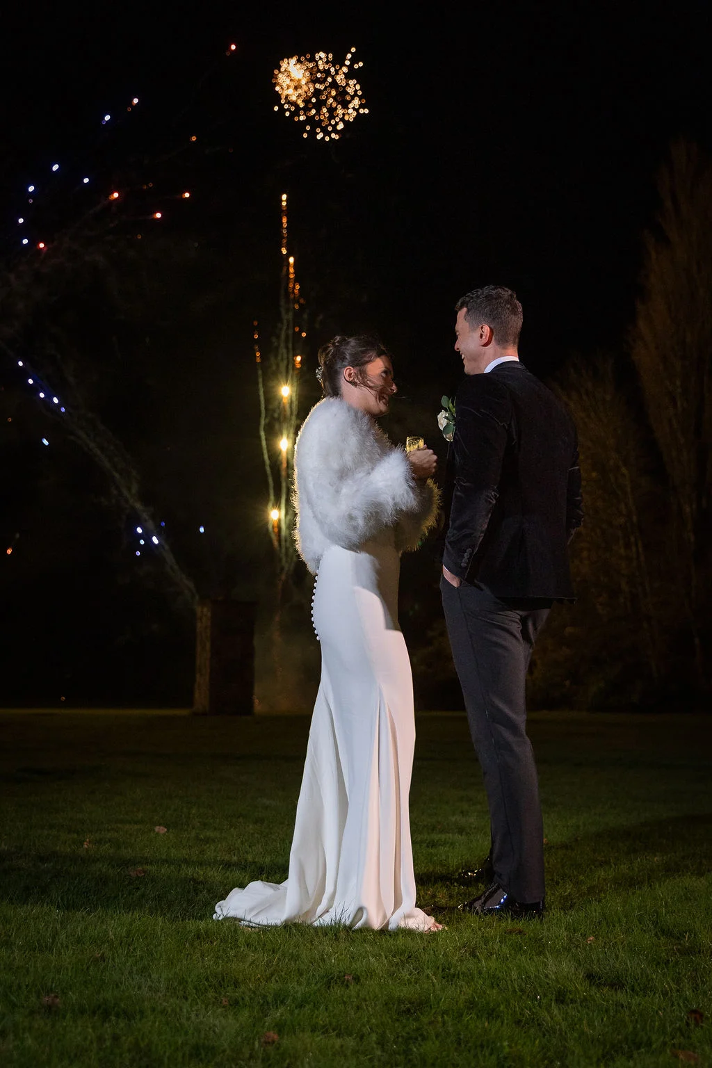 A bride and groom laugh together on a lawn at night, framed by a spectacular fireworks display lighting up the dark sky.