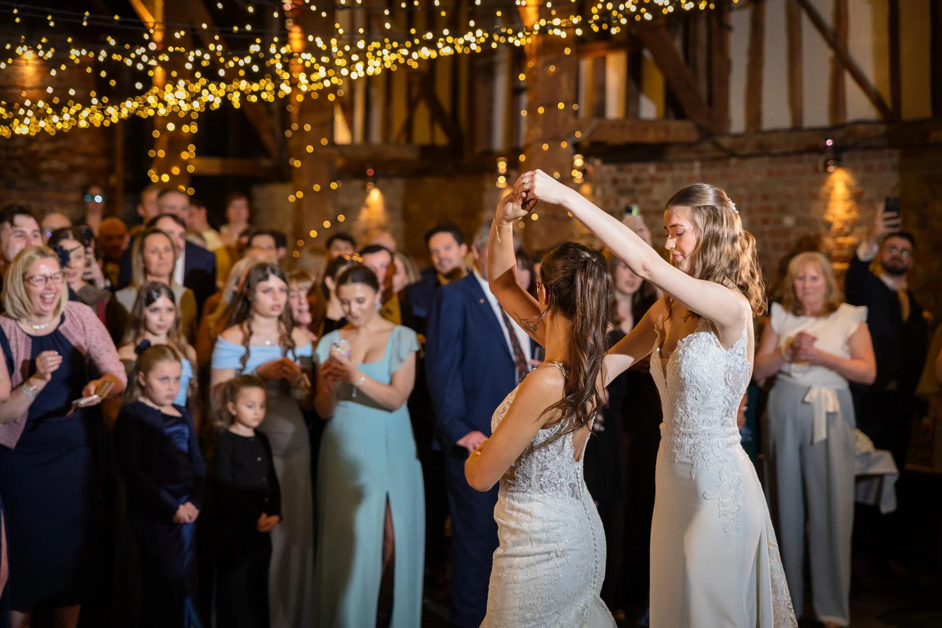 Two brides share their first dance under a magical canopy of twinkling fairy lights inside a rustic oak-framed barn, surrounded by smiling wedding guests.