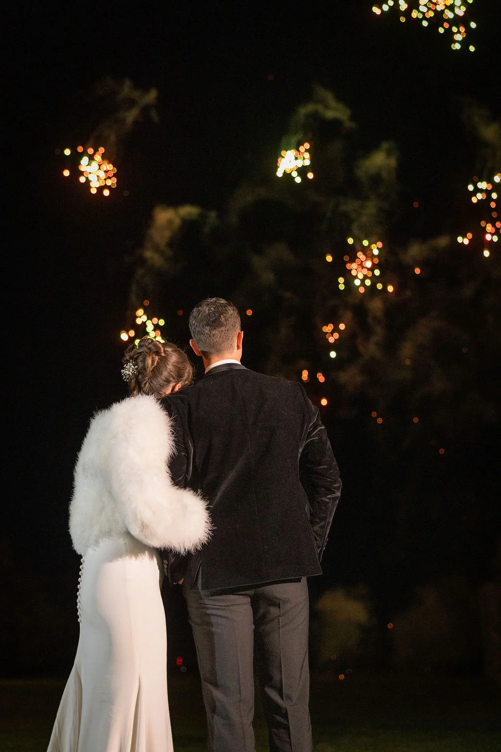 A newlywed couple viewed from behind watch a spectacular fireworks display lighting up the night sky during their winter wedding reception.