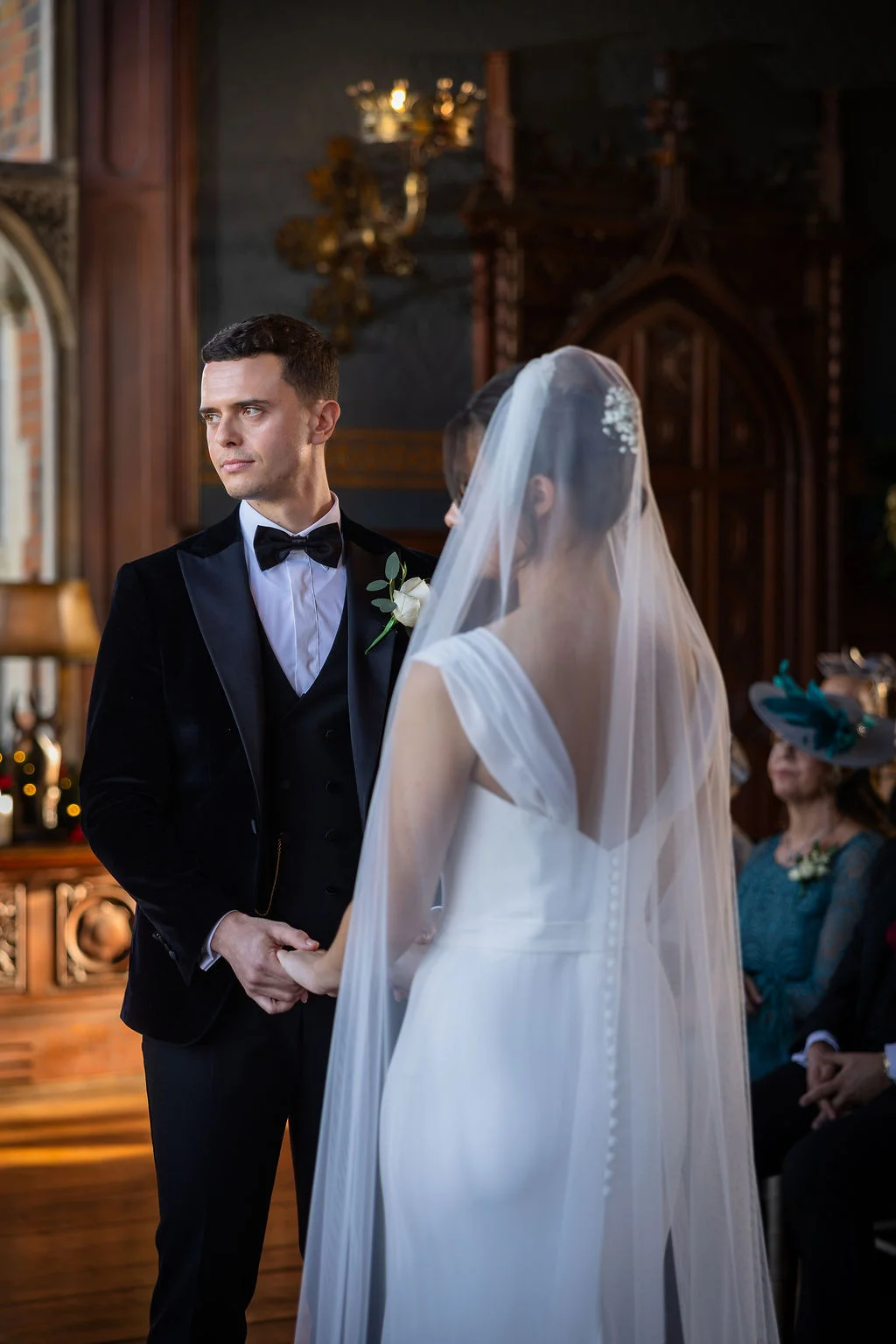 A groom in a black velvet tuxedo looks away thoughtfully while holding hands with his bride during their ceremony inside a historic venue.