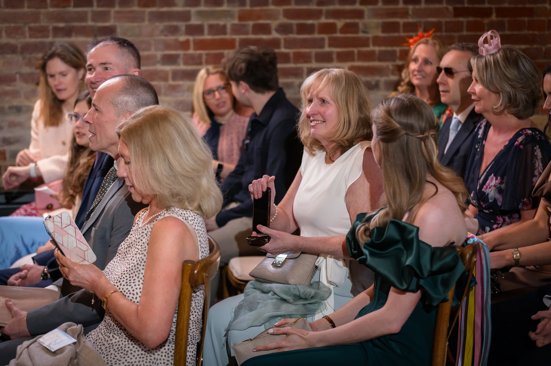 A candid shot of wedding guests seated indoors against an exposed brick wall, watching the ceremony unfold with attentive expressions.