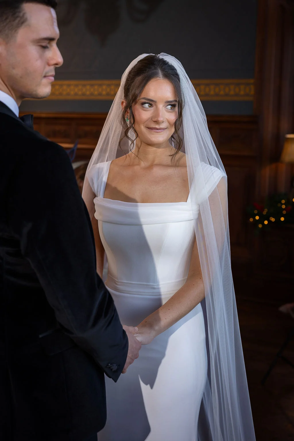 The bride and groom hold hands during an intimate ceremony inside a wood-panelled manor house, with the bride wearing a sleek square-neck gown and veil.
