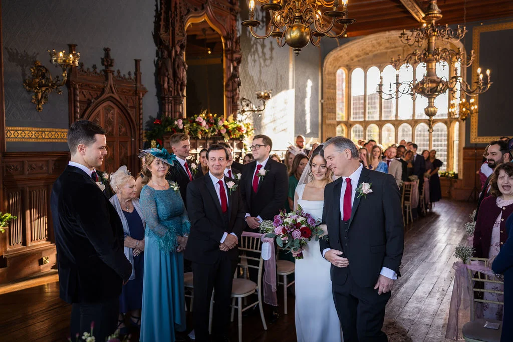 A bride walks down the aisle accompanied by her father in a grand, wood-panelled historic hall filled with guests during a traditional wedding ceremony.