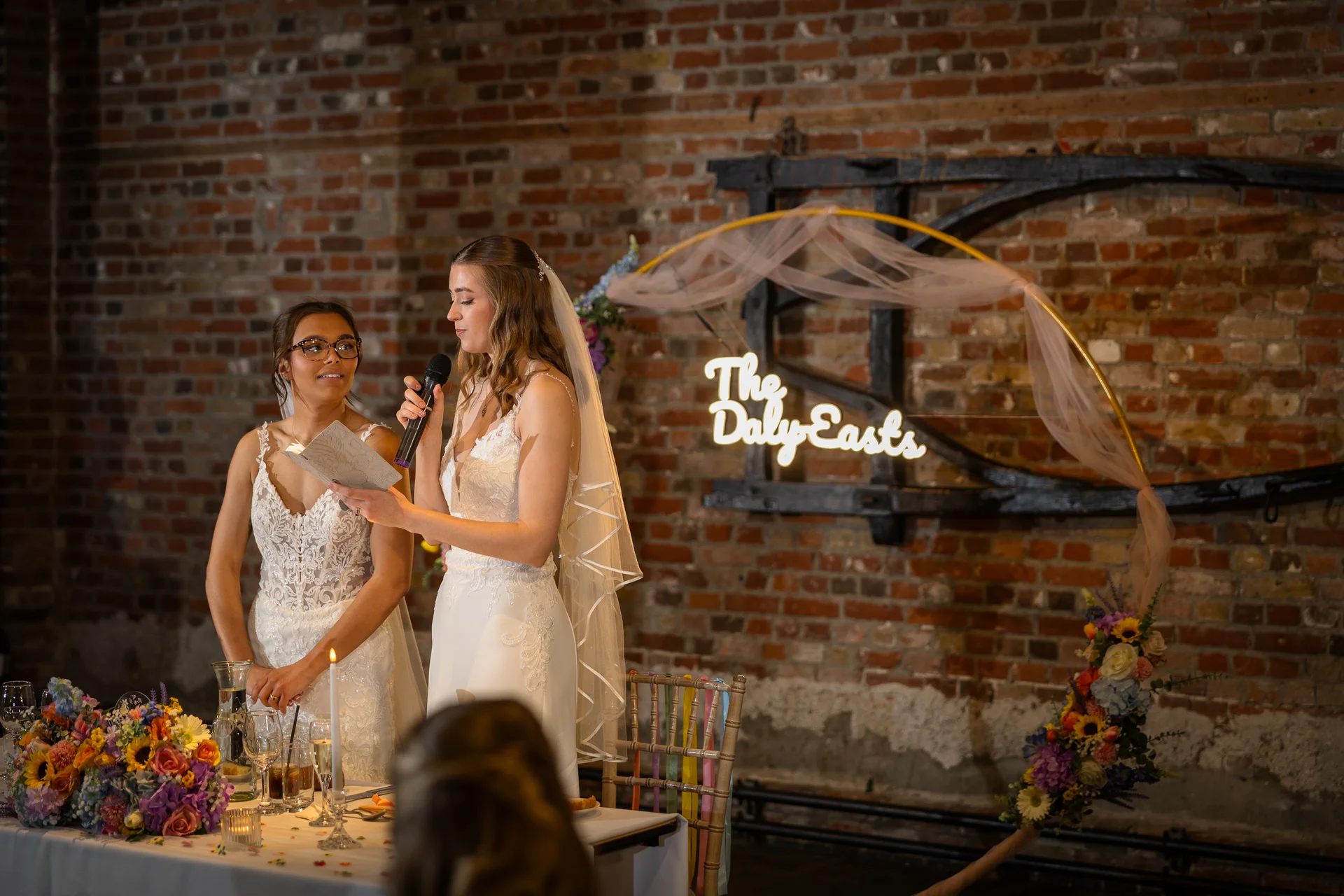 A bride gives a heartfelt speech holding a microphone next to her bridesmaid in an exposed brick venue decorated with a neon sign and floral arch.