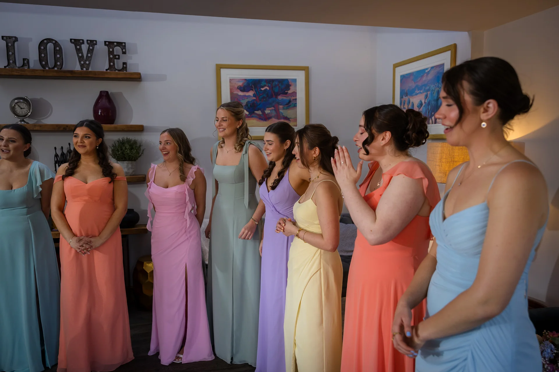 A group of bridesmaids wearing colourful, mismatched pastel dresses stand together indoors, smiling and reacting to a moment.