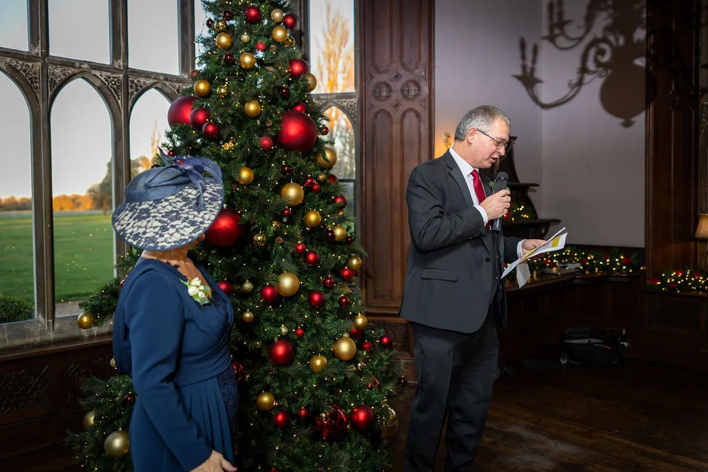 A father of the bride delivers a speech holding a microphone and papers near a large decorated Christmas tree in a historic country house venue.