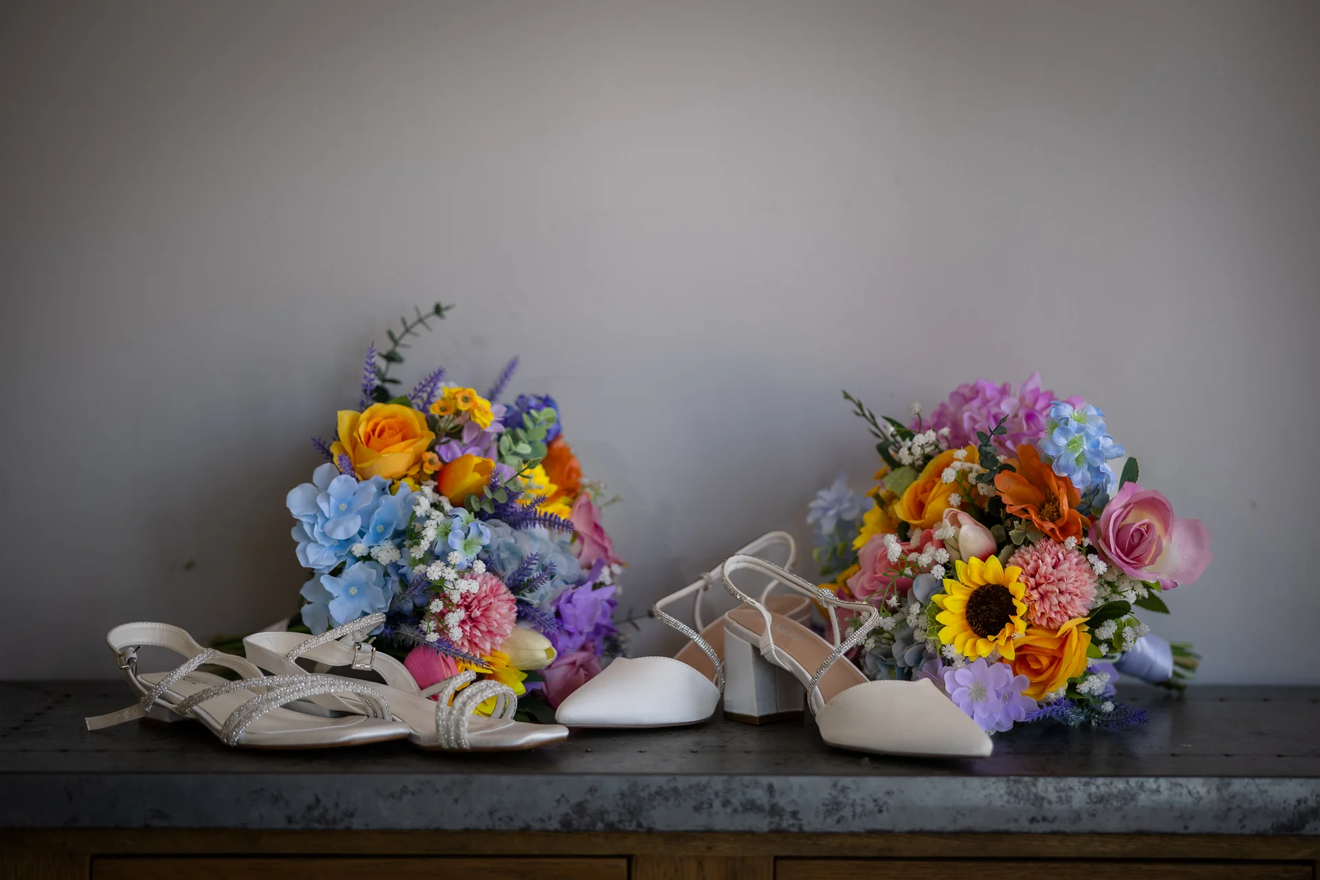 Two pairs of elegant white bridal shoes are displayed alongside vibrant, colourful wedding bouquets featuring roses, sunflowers, and hydrangeas on a dark surface.