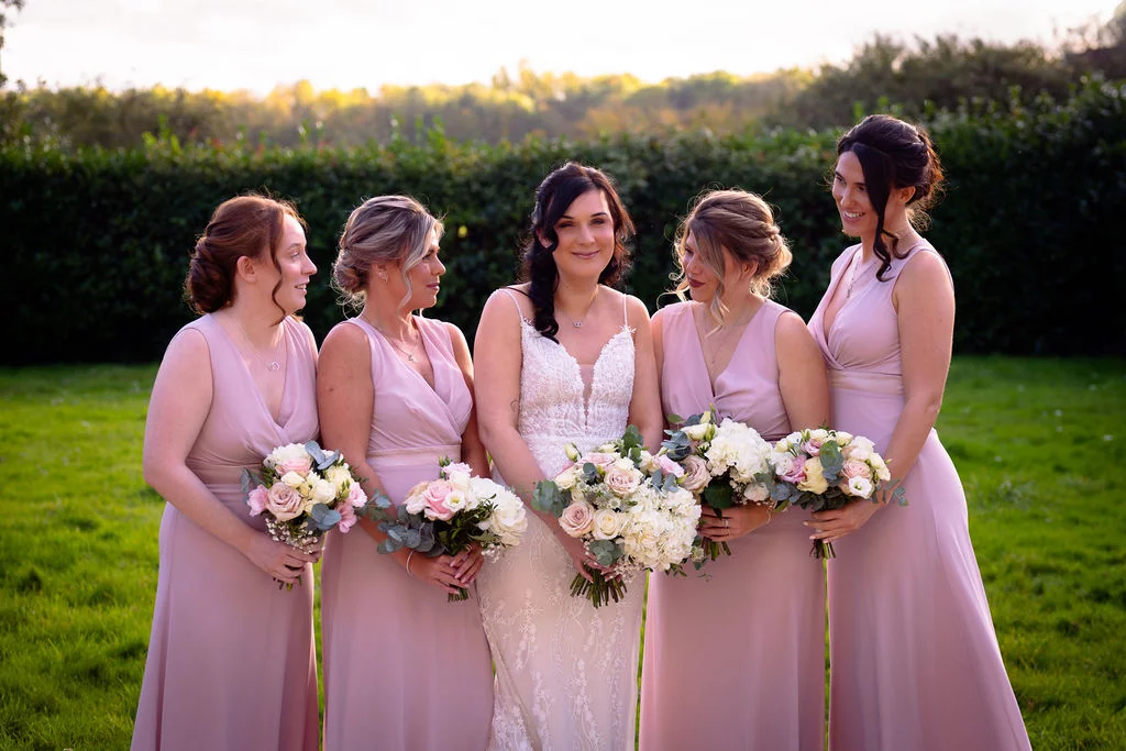 A smiling bride stands outdoors on a lawn at golden hour flanked by four bridesmaids in matching pink dresses holding bouquets of roses and greenery.