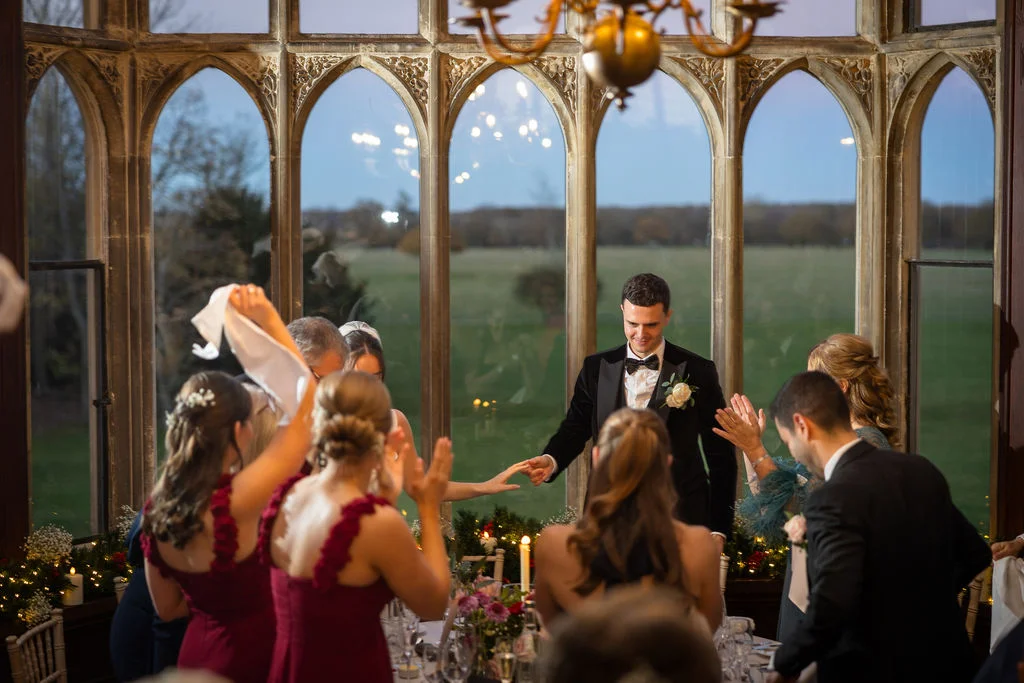 A groom in a velvet tuxedo stands smiling during wedding speeches inside a grand historic reception room with large arched windows at dusk.