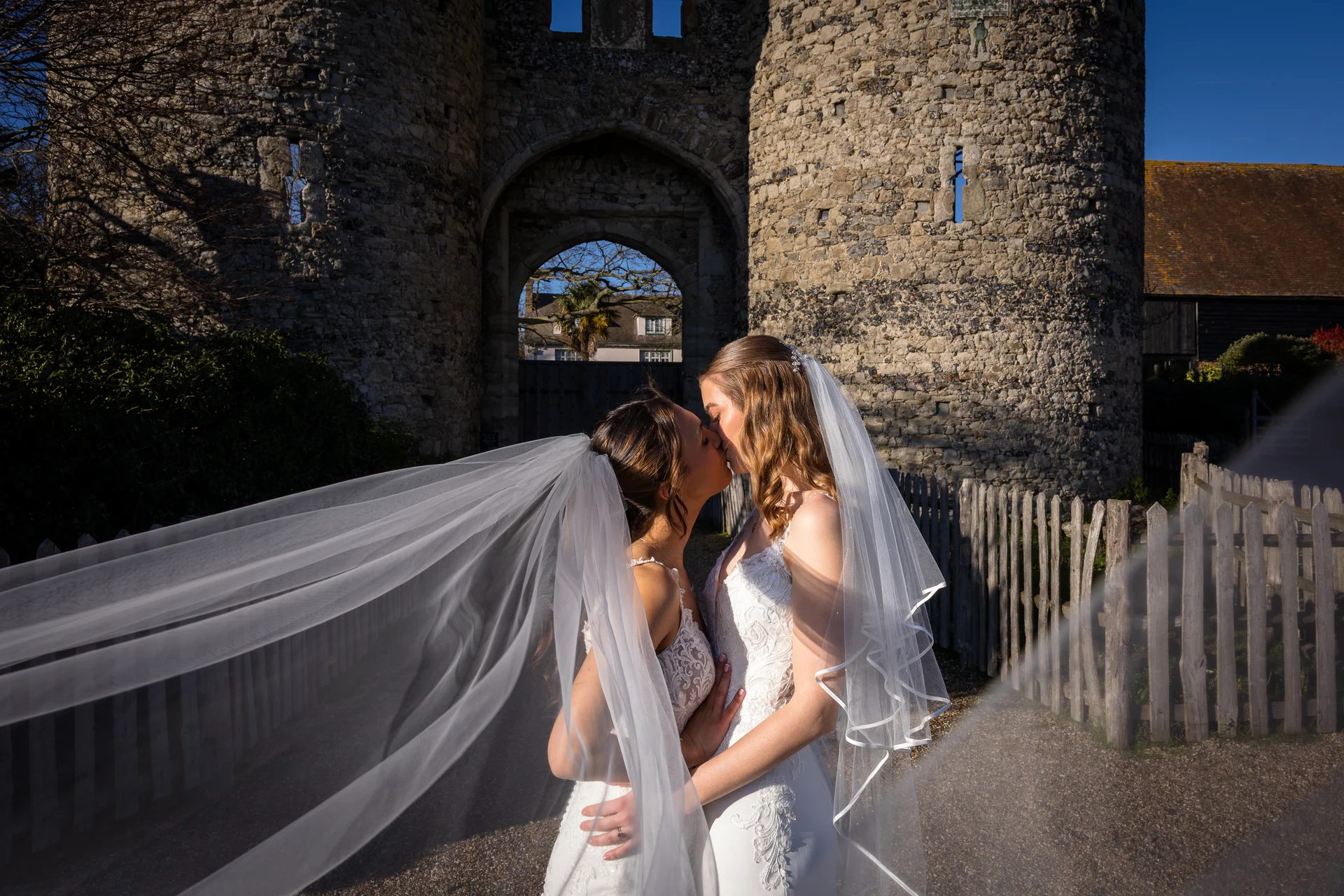 Two brides share a tender kiss in front of a historic stone castle gateway as their veils blow dramatically in the wind.