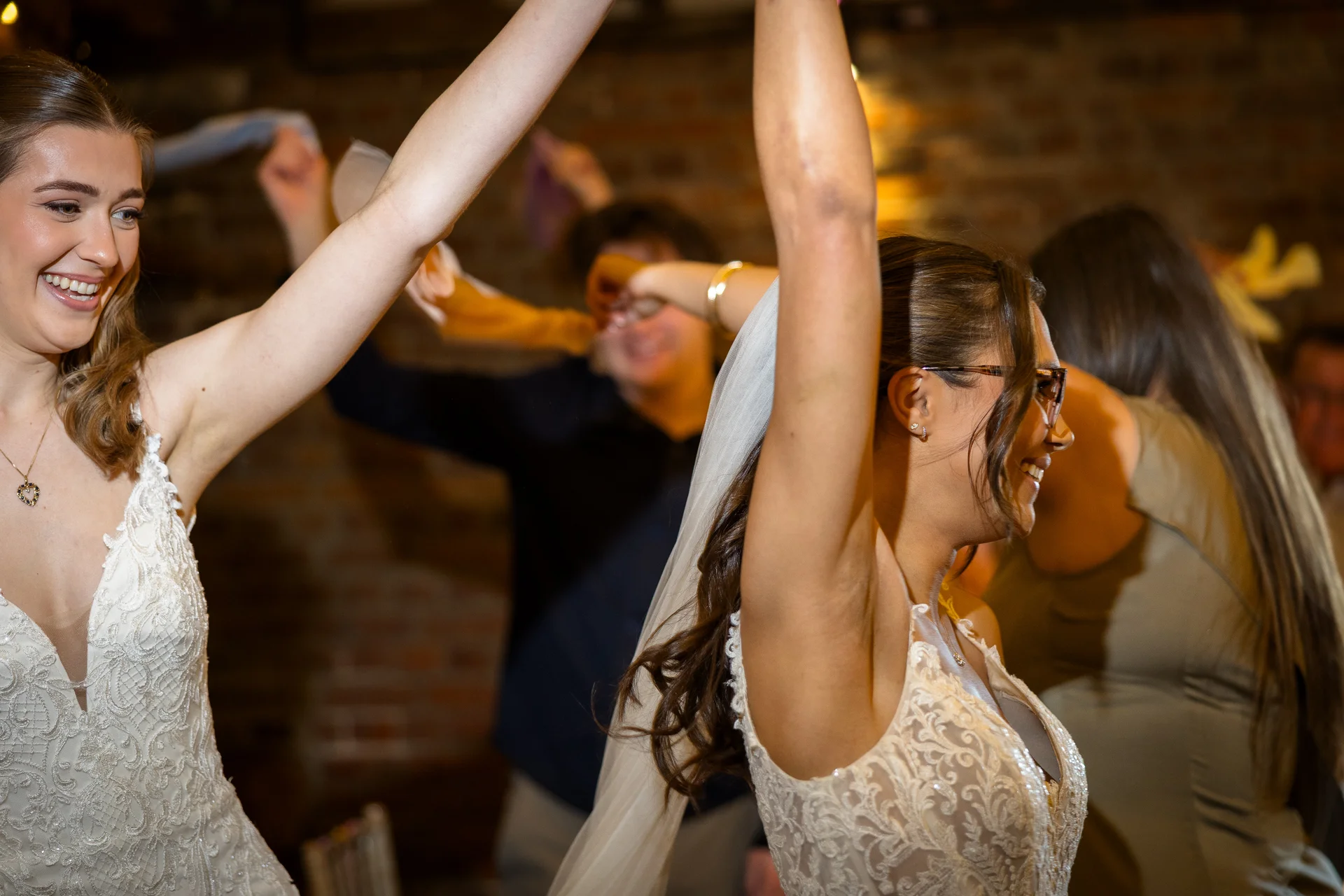 Two smiling brides with arms raised and veils flowing are captured dancing joyfully in a rustic venue with exposed brick walls.