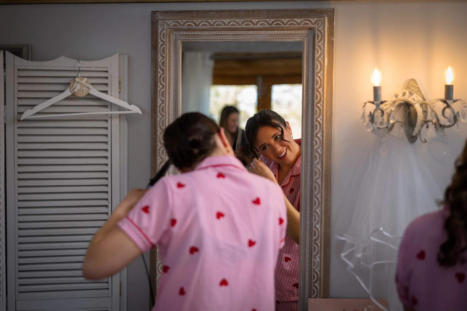 A smiling bride styling her hair in front of a ornate mirror while wearing pink patterned pyjamas during morning bridal preparations.