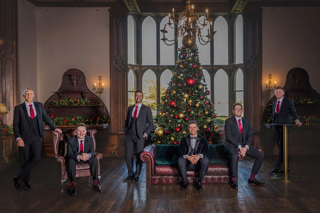 A groom and his groomsmen pose together in a grand, wood-panelled stately home featuring a large decorated Christmas tree and leather furniture.