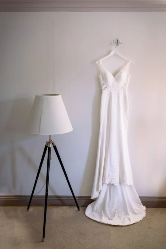A elegant lace and crepe wedding dress hangs neatly against a plain wall next to a modern tripod floor lamp during morning preparations.