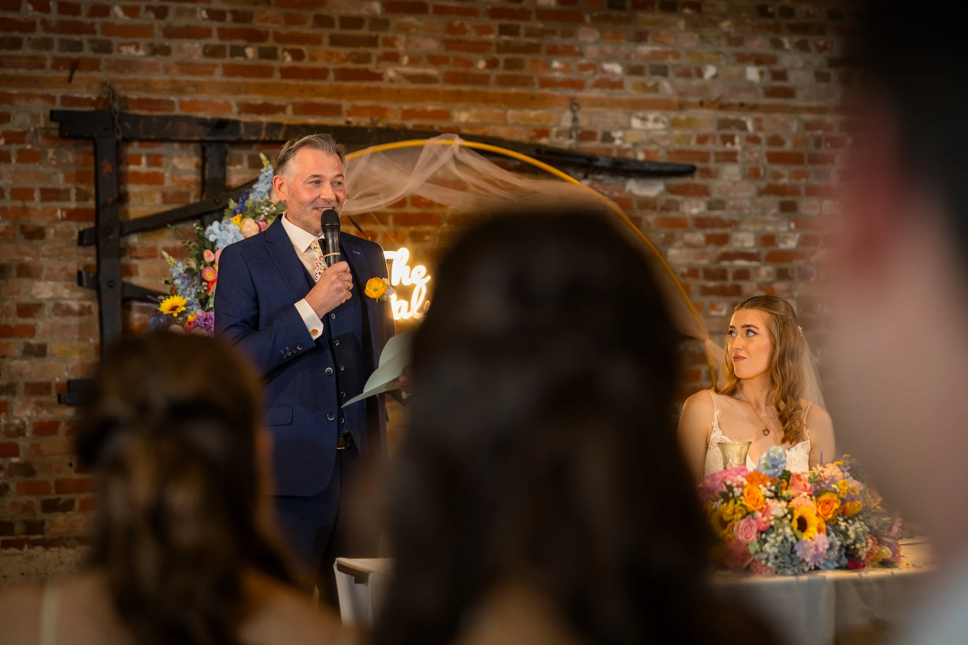 A father in a navy suit happily delivers a wedding speech with a microphone, while a bride looks on admiringly in front of a rustic brick wall.