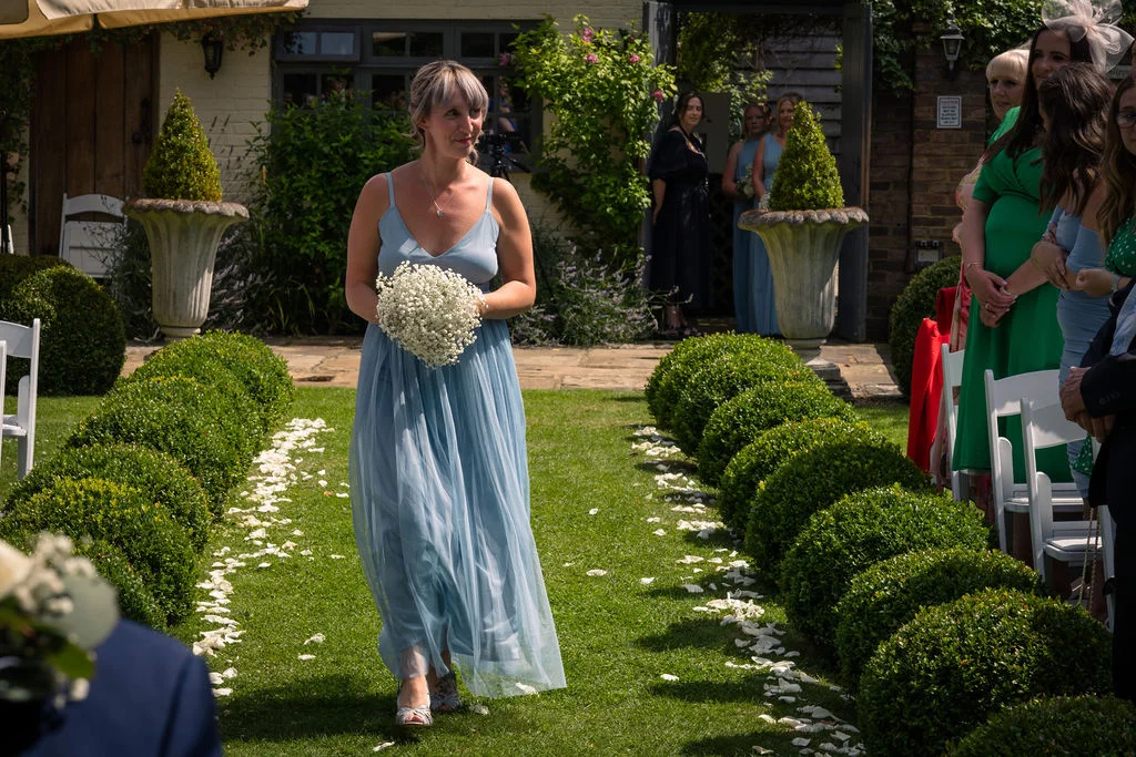 A bridesmaid in a dusty blue dress and carrying a baby's breath bouquet walks down a grass aisle lined with topiary bushes and scattered petals.
