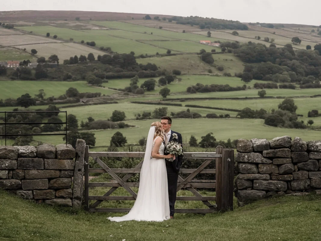 A bride and groom standing in front of a stone gate in the countryside.