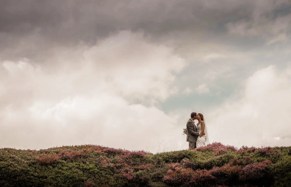 A bride and groom standing on top of a hill under a cloudy sky.
