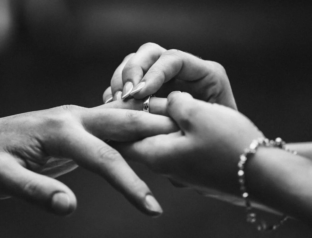 A black and white photo of a couple putting a wedding ring on each other's hands.