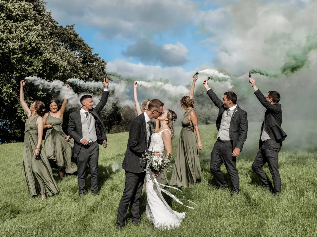A group of bridesmaids and groomsmen in a field with green smoke.