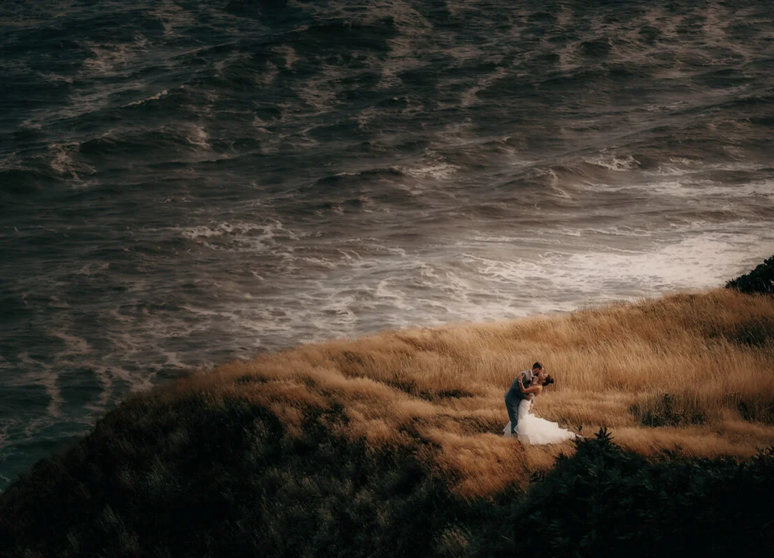 A bride and groom standing on a grassy hill overlooking the ocean.