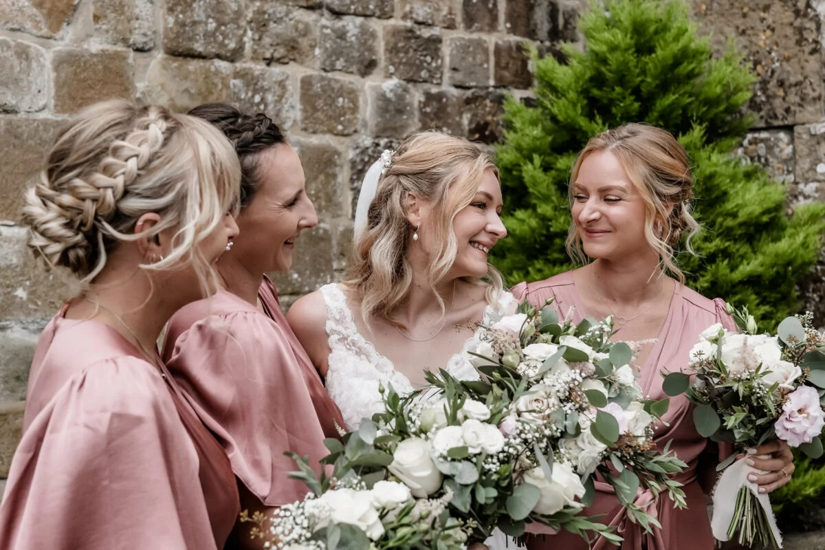 Bridesmaids in pink robes with bouquets in front of a stone wall.