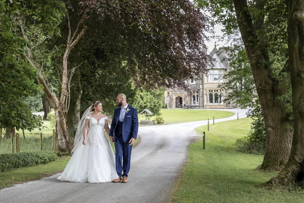A bride and groom walking down a path in front of a large mansion.