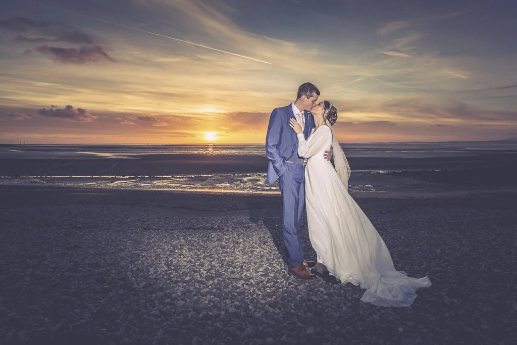 A bride and groom kissing on the beach at sunset.
