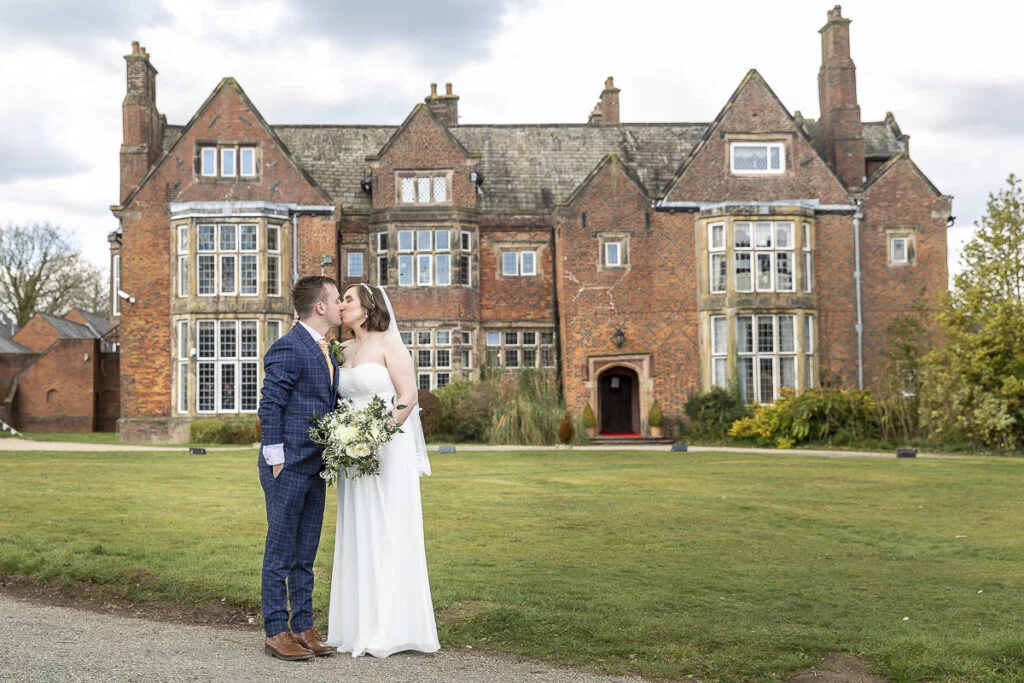 A bride and groom kissing in front of a large mansion.