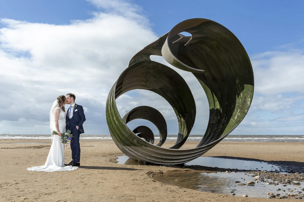 A bride and groom standing next to a sculpture on the beach.