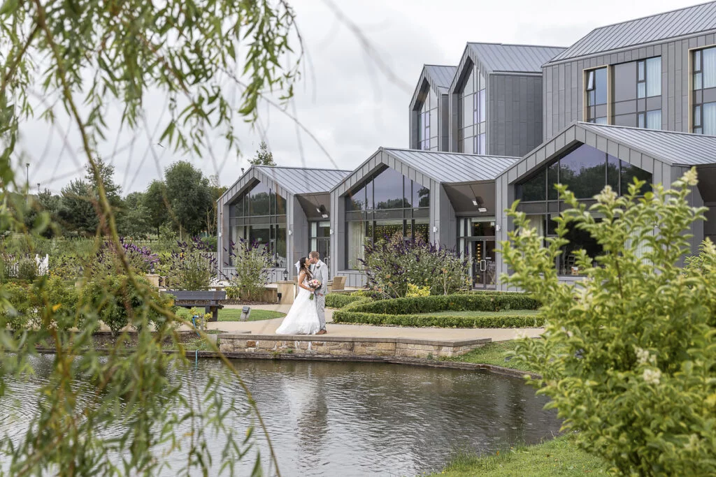 A bride and groom standing in front of a building with a pond.