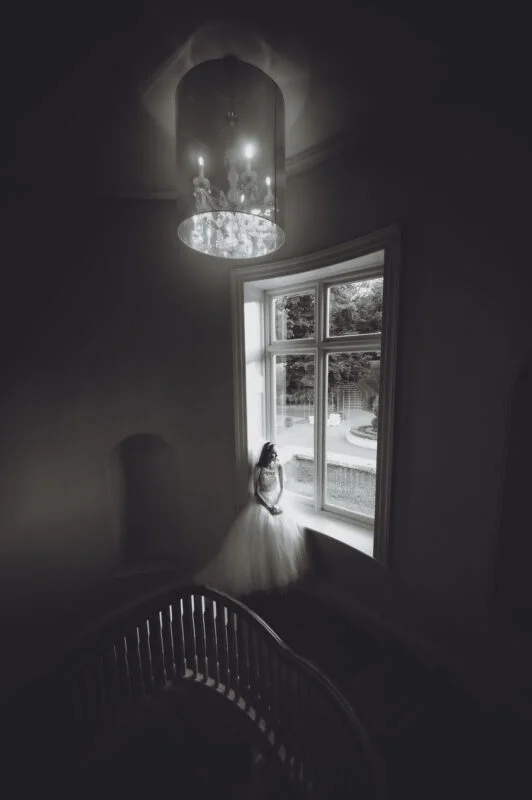 A black and white photo of a girl in a wedding dress looking out of a window.