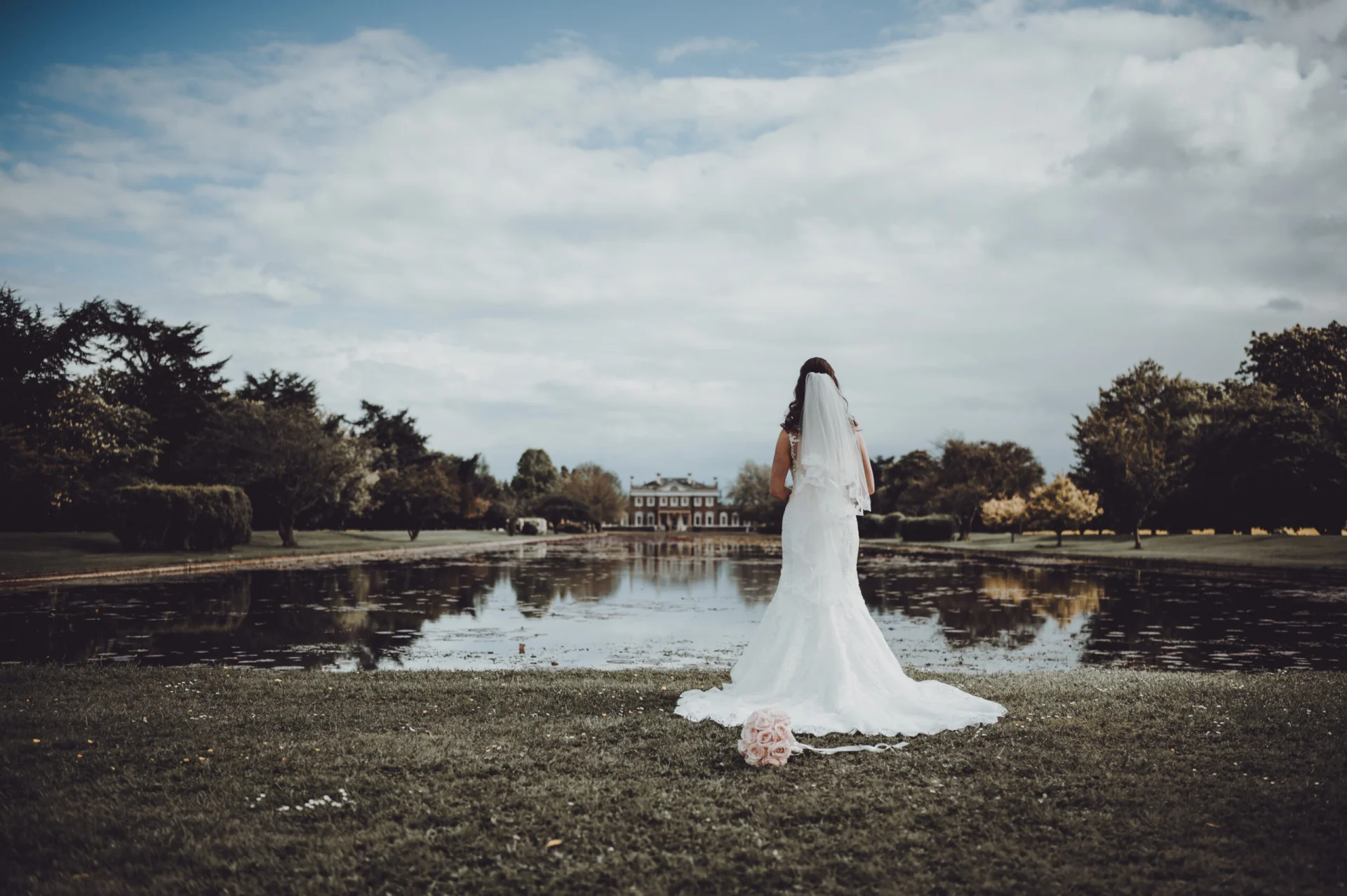 A bride in a lace fit and flare gown stands with her back to the camera by a tranquil lakeside with a grand manor house in the distance.